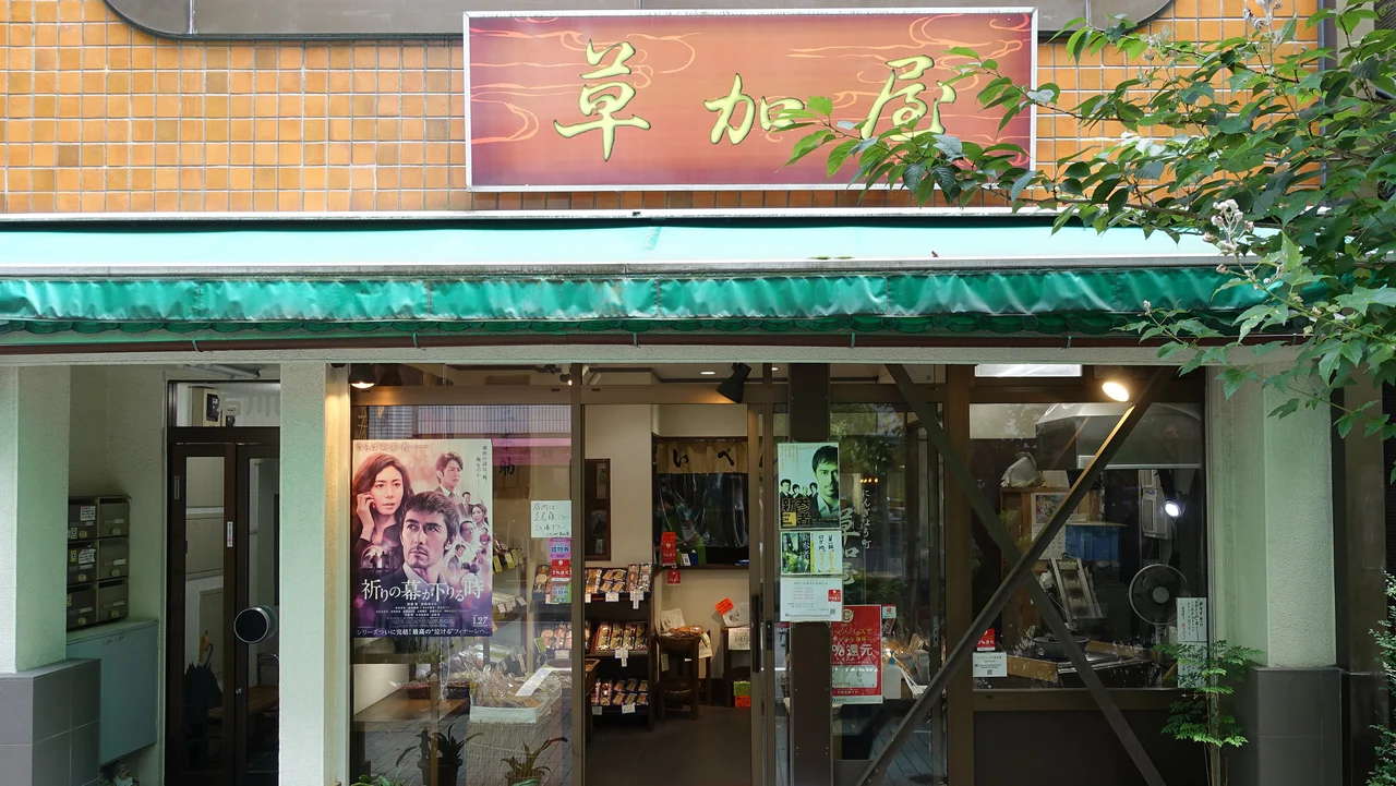 草加屋の外観。『麒麟の翼』のポスターが貼られた店先には、煎餅や和菓子が並ぶ。東野圭吾ファンの巡礼地としても知られる。